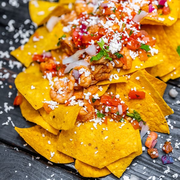 Shrimp nachos with pico de gallo and crumbled white cheese on a dark wooden surface.