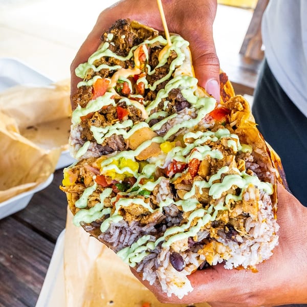 Hands holding a large, cut-open Tiagos Tacos burrito filled with meat, rice, vegetables, and green sauce.
