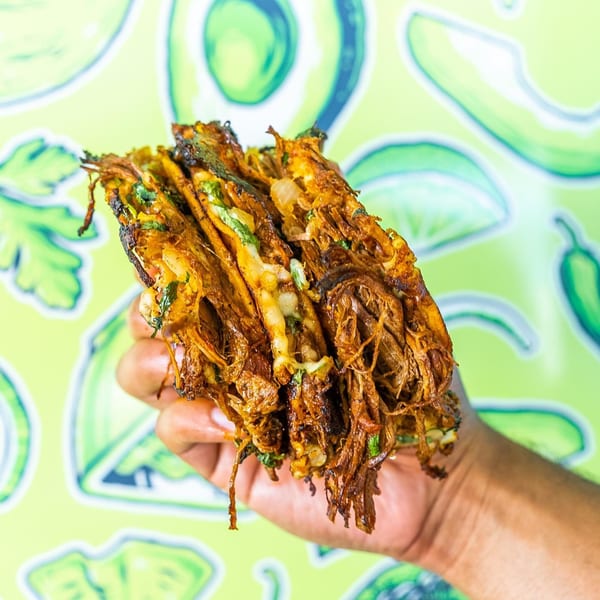 Hand holding a stack of crispy, cheesy Tiagos Tacos with shredded meat against an avocado-patterned wall.