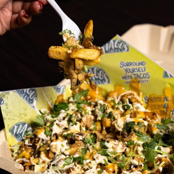 A fork lifts seasoned fries and meat from a Tiagos Tacos container with cilantro and sauces.