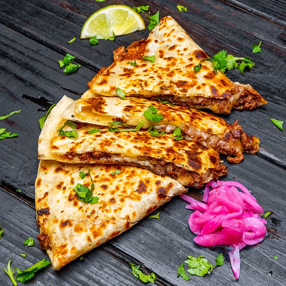 Three Tiagos Tacos quesadillas with meat and cheese, garnished with cilantro, lime, and pickled onions.