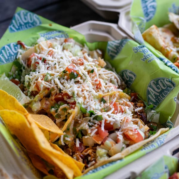 Tiagos Tacos take-out box filled with tacos, topped with cheese, tomatoes, and cilantro, with tortilla chips.