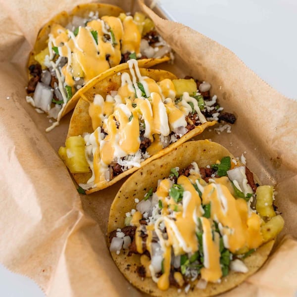 Three Tiagos Tacos with meat, pineapple, onions, cilantro, and two sauces in a paper-lined tray.