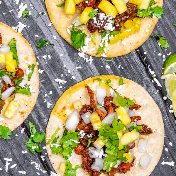 Three Tiagos Tacos with meat, pineapple, onions, cilantro, and cheese on a dark wooden surface.