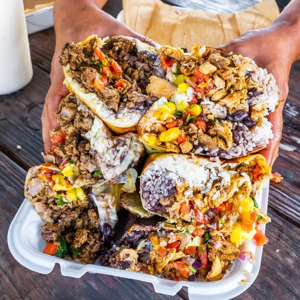 Hands holding a large takeout container overflowing with several Tiagos Tacos burritos, filled with meat, rice, and salsa.